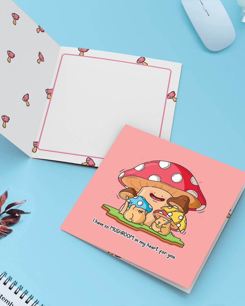 Stylish flat lay showing the mushroom pun card open on a pastel blue desk with a calendar and yellow flowers. Designed by Toastedink and stocked at Rebel Romance, this square card features adorable mushroom illustrations and space to write a personal message inside.