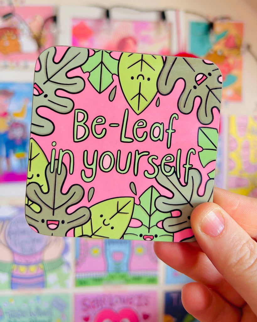 A hand holding the Rebel Romance “Be‑Leaf In Yourself” coaster by Kelly Lou. Featuring smiling green leaves on a pink background, this glossy square coaster is a fun, positive homeware accessory from a UK indie brand for alternative aesthetics.