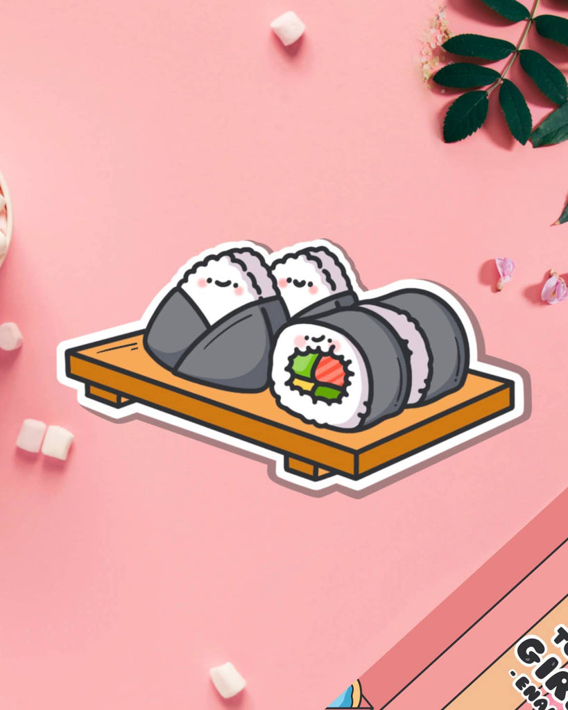 Cute vinyl sticker of a sushi platter featuring happy sushi rolls and onigiri with smiling faces, placed on a wooden tray. Photographed on a pink background with mini marshmallows and floral elements. Sold by Rebel Romance, a UK brand celebrating alternative style and quirky accessories.