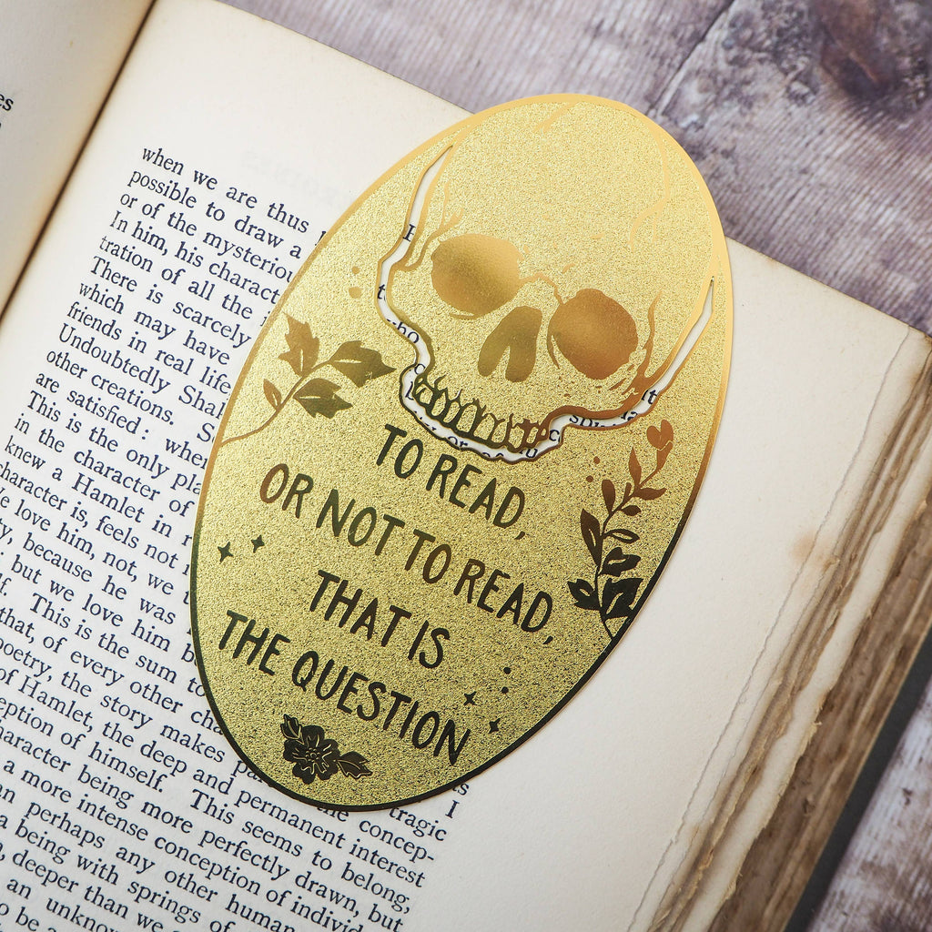 A close-up of a brass bookmark shaped like a skull, elegantly perched at the top of an open book. The intricate design of the skull is etched into the brass, giving it a striking and detailed appearance. The book’s page is titled “Ophelia,” referencing Shakespeare’s Hamlet.