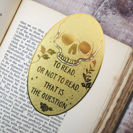 A close-up of a brass bookmark shaped like a skull, elegantly perched at the top of an open book. The intricate design of the skull is etched into the brass, giving it a striking and detailed appearance. The book’s page is titled “Ophelia,” referencing Shakespeare’s Hamlet.
