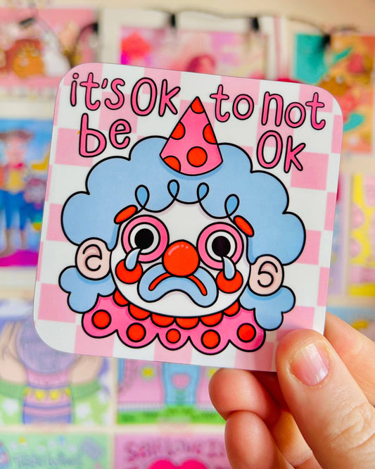 A person holding a glossy Rebel Romance coaster designed by Kellylou, showing a sad clown with blue hair, a red nose, and big tears, with the phrase "it's OK to not be OK" above. The background is a pink checkerboard. The scene is set against a vibrant display of cute and colourful artwork.
