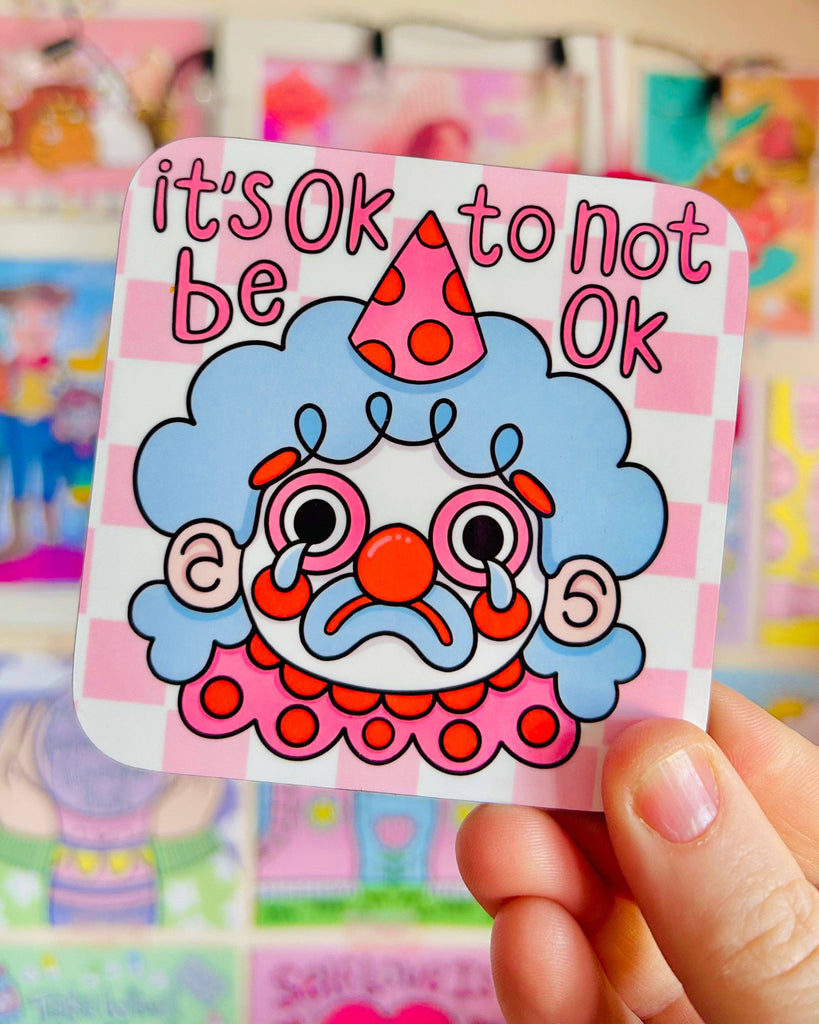A person holding a glossy Rebel Romance coaster designed by Kellylou, showing a sad clown with blue hair, a red nose, and big tears, with the phrase "it's OK to not be OK" above. The background is a pink checkerboard. The scene is set against a vibrant display of cute and colourful artwork.