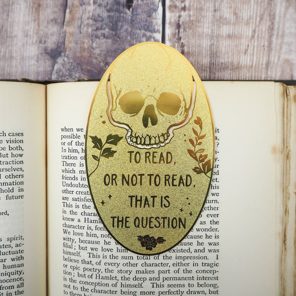A close-up of a brass bookmark shaped like a skull, elegantly perched at the top of an open book. The intricate design of the skull is etched into the brass, giving it a striking and detailed appearance. The book’s page is titled “Ophelia,” referencing Shakespeare’s Hamlet.