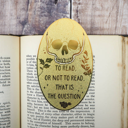 A close-up of a brass bookmark shaped like a skull, elegantly perched at the top of an open book. The intricate design of the skull is etched into the brass, giving it a striking and detailed appearance. The book’s page is titled “Ophelia,” referencing Shakespeare’s Hamlet.