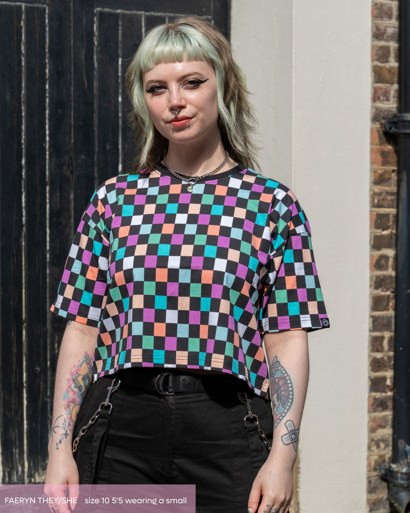 Faeryn, a non binary femme person wearing a colourblock checkerboard crop top with purple, orange, green, blue and pastel tones with black and a black contrasting neckline. They've paired it with black oversized trousers. They're stood smiling with their hands resting by their side with a black door behind them. 