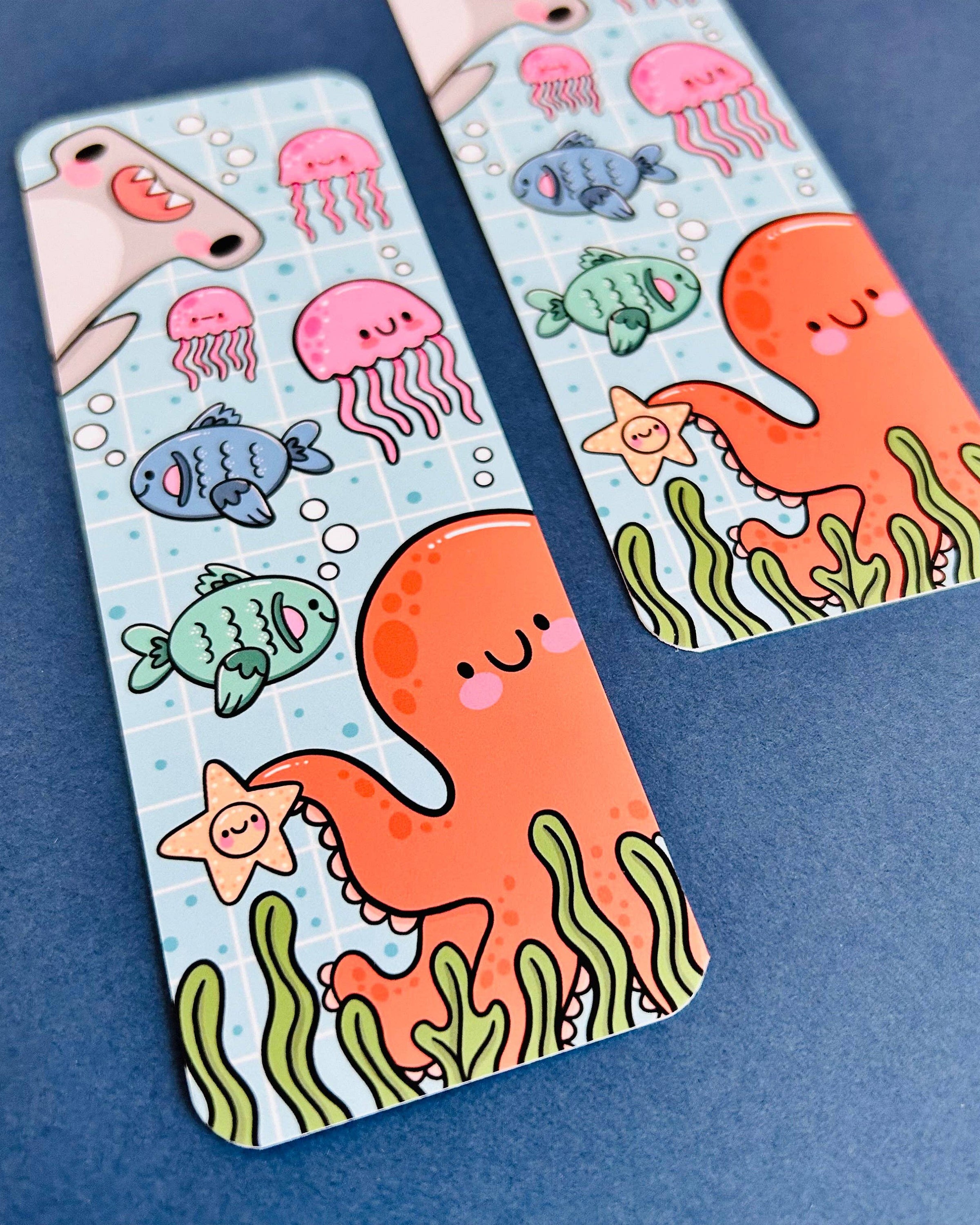 Close-up of two Rebel Romance Under The Sea bookmarks designed by Kelly Lou, featuring a cute orange octopus holding a starfish, surrounded by jellyfish, smiling fish, seaweed, and a shark on a blue grid background. Printed on velvet-laminated card with rounded corners, perfect for adding ocean-themed joy to your reading. Displayed on a blue surface for a bright and playful look.