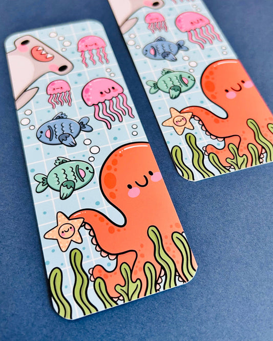 Close-up of two Rebel Romance Under The Sea bookmarks designed by Kelly Lou, featuring a cute orange octopus holding a starfish, surrounded by jellyfish, smiling fish, seaweed, and a shark on a blue grid background. Printed on velvet-laminated card with rounded corners, perfect for adding ocean-themed joy to your reading. Displayed on a blue surface for a bright and playful look.