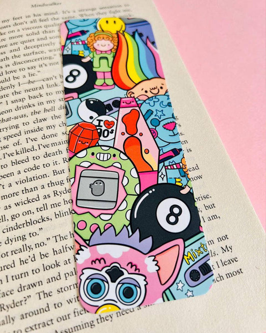 Colourful 90s Nostalgia Bookmark by Kelly Lou from Rebel Romance, placed on top of an open book. The velvet-laminated design features retro icons like a lava lamp, troll doll, Furby, Tamagotchi, and 8-ball in a bold, playful cartoon style. A perfect gift for millennials who love cute, nostalgic stationery from this small UK business.
