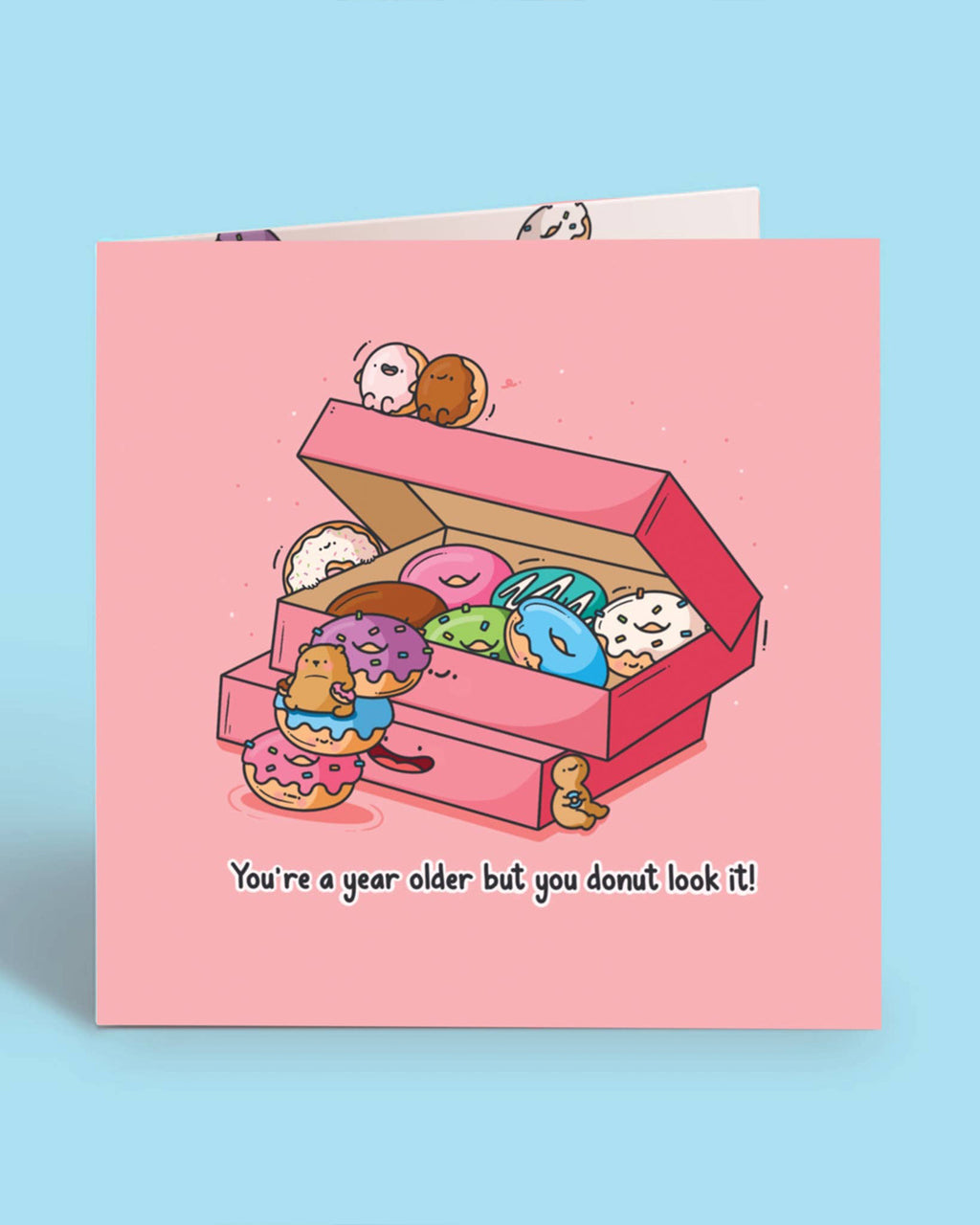 Front cover of the Toastedink birthday card featuring a pink box of colourful cartoon donuts with smiling faces and a bear climbing the donuts, with the caption 