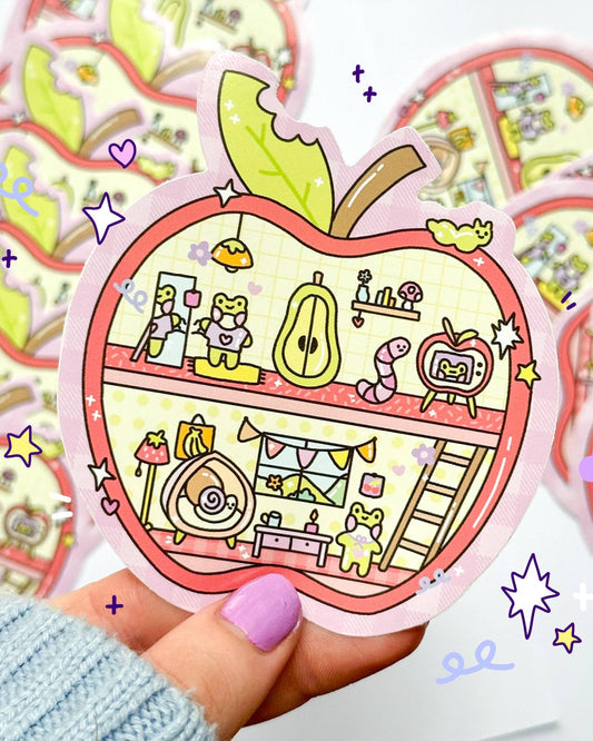 Cute Apple House vinyl sticker by Sun Puff Studios, sold by Rebel Romance. The design features an adorable cross-section of an apple-shaped home with pastel characters like a frog, worm, snail, and retro TV. Illustrated in 90s toy-inspired sketch style with soft colours. Perfect for scrapbooking, journalling, or decorating laptops and water bottles. Waterproof and glossy finish. Great for trinket lovers and stationery collectors.
