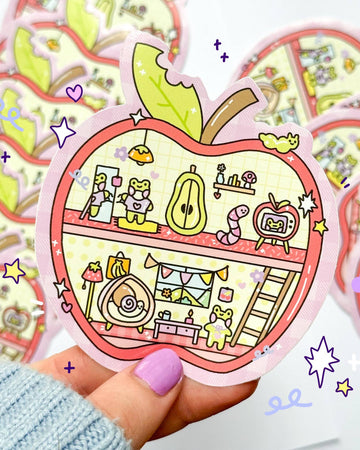 Cute Apple House vinyl sticker by Sun Puff Studios, sold by Rebel Romance. The design features an adorable cross-section of an apple-shaped home with pastel characters like a frog, worm, snail, and retro TV. Illustrated in 90s toy-inspired sketch style with soft colours. Perfect for scrapbooking, journalling, or decorating laptops and water bottles. Waterproof and glossy finish. Great for trinket lovers and stationery collectors.