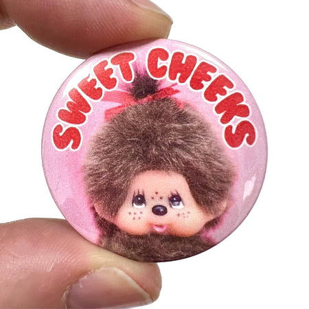 A 38mm high-gloss pin badge featuring a retro-style monkey with fluffy fur and a cheeky expression. The text “Sweet Cheeks” is written in bold red letters on a pink background. Designed by Bite Your Granny.