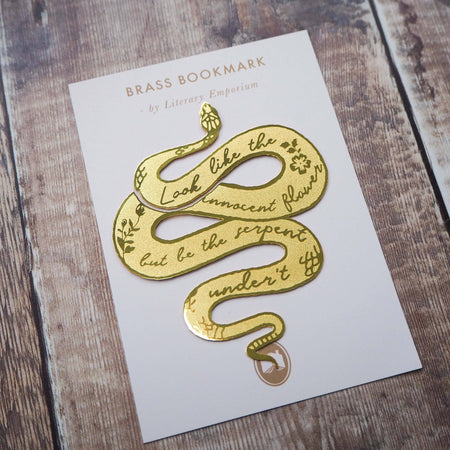 A brass bookmark shaped like a coiled serpent, delicately etched with the famous Lady Macbeth quote: “Look like the innocent flower, But be the serpent under ‘t.” The bookmark is placed inside an open book on a wooden surface, with the title “Lady Macbeth” visible on the page. The snake’s head is designed to peek out from the top of the book when closed, adding a dramatic and literary flair.