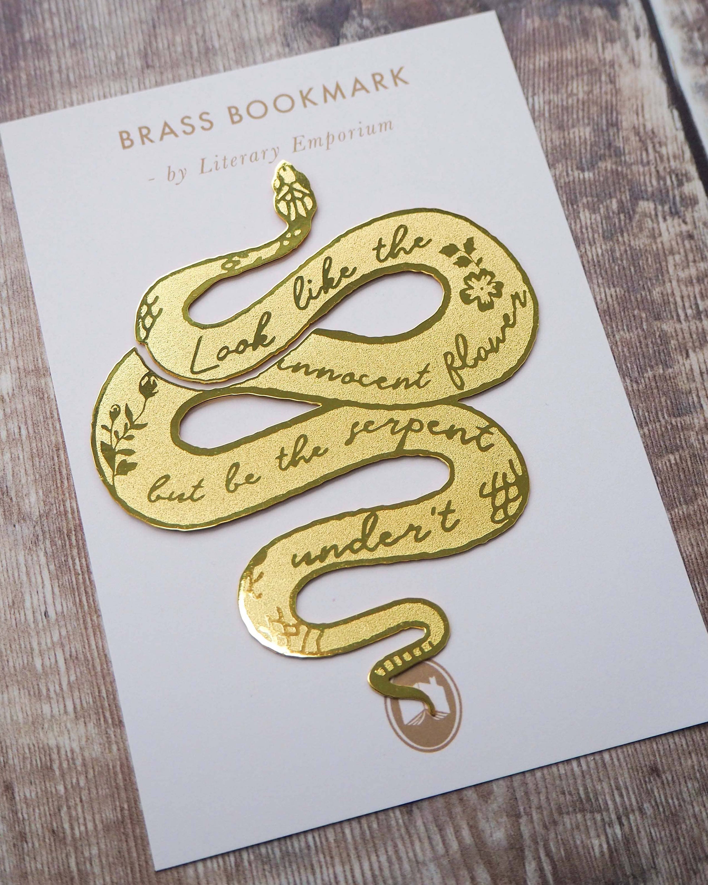 A brass bookmark shaped like a coiled serpent, delicately etched with the famous Lady Macbeth quote: “Look like the innocent flower, But be the serpent under ‘t.” The bookmark is placed inside an open book on a wooden surface, with the title “Lady Macbeth” visible on the page. The snake’s head is designed to peek out from the top of the book when closed, adding a dramatic and literary flair.