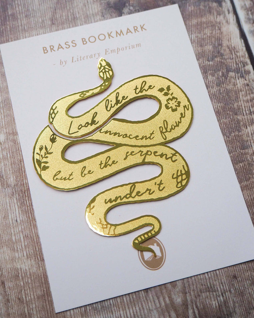 A brass bookmark shaped like a coiled serpent, delicately etched with the famous Lady Macbeth quote: “Look like the innocent flower, But be the serpent under ‘t.” The bookmark is placed inside an open book on a wooden surface, with the title “Lady Macbeth” visible on the page. The snake’s head is designed to peek out from the top of the book when closed, adding a dramatic and literary flair.