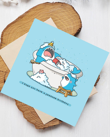 Funny shark birthday card by Toastedink, sold by Rebel Romance. Shows a cartoon shark relaxing in a bubble bath with a book, while a smaller shark stands on a stool. Blue card with the pun "I hope you have a jawsome birthday." Includes envelope.
