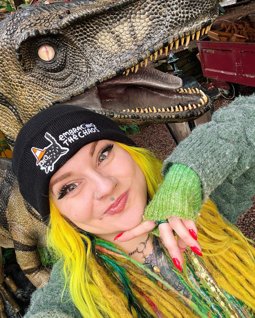 Katie Abey wearing the black Embracing The Chaos Cat Beanie Hat by Katie Abey standing in front of a dinosaur sculpture. She has bright yellow hair and colourful accessories. The beanie shows the embroidered chaotic cat and text Embracing The Chaos. Cosy ethical winter hat for quirky style lovers.