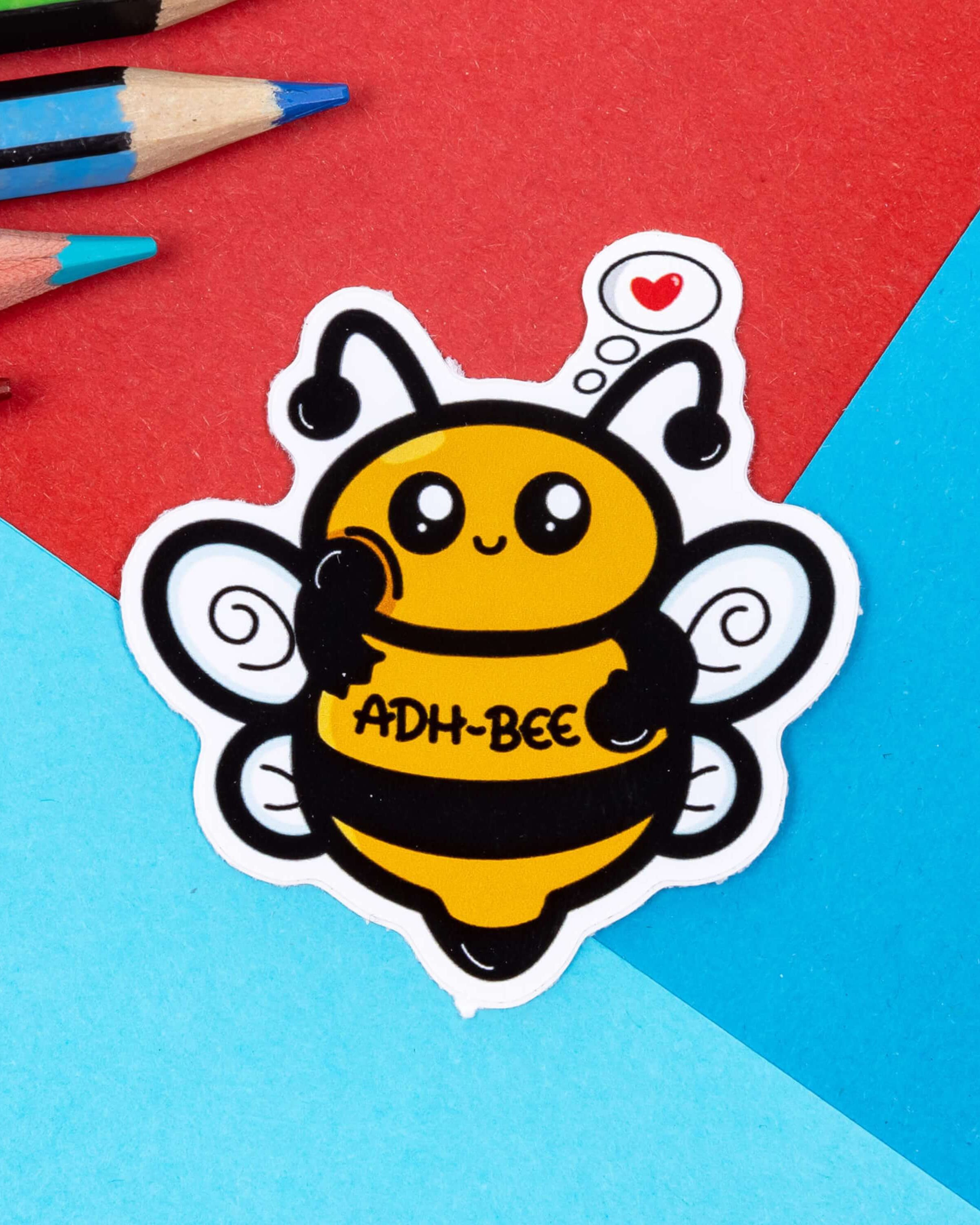Cute ADH Bee sticker by Innabox, available at Rebel Romance. This adorable bee-shaped vinyl sticker features a happy yellow and black cartoon bee with swirly wings, a love heart thought bubble, and the words 