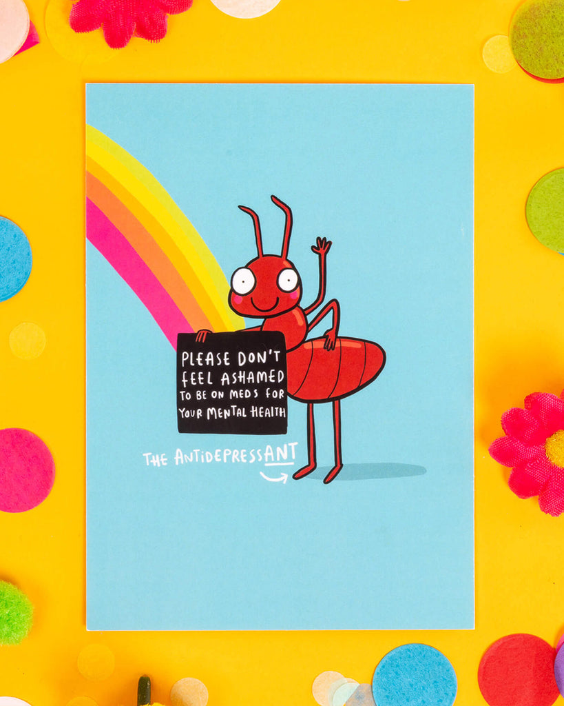 A light blue A6 postcard featuring a red cartoon ant holding a sign that says “Please don’t feel ashamed to be on meds for your mental health,” with the caption “The Antidepressant.” A rainbow streams behind the ant. Designed by Katie Abey, this quirky mental health postcard is ideal for millennials into alternative humour, self-care, and spreading destigmatising messages with colourful stationery.