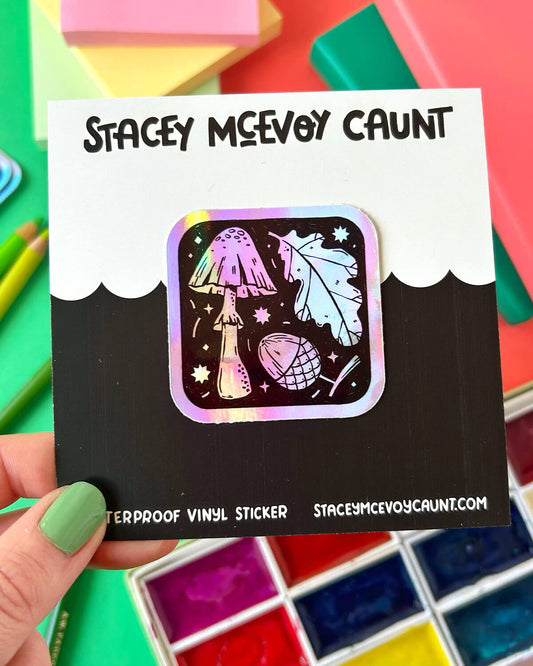 A square rainbow holographic sticker with illustrations of a mushroom, oak leaf, and acorn on a starry black background. The sticker shimmers with rainbow hues, held by a hand with green nail polish.