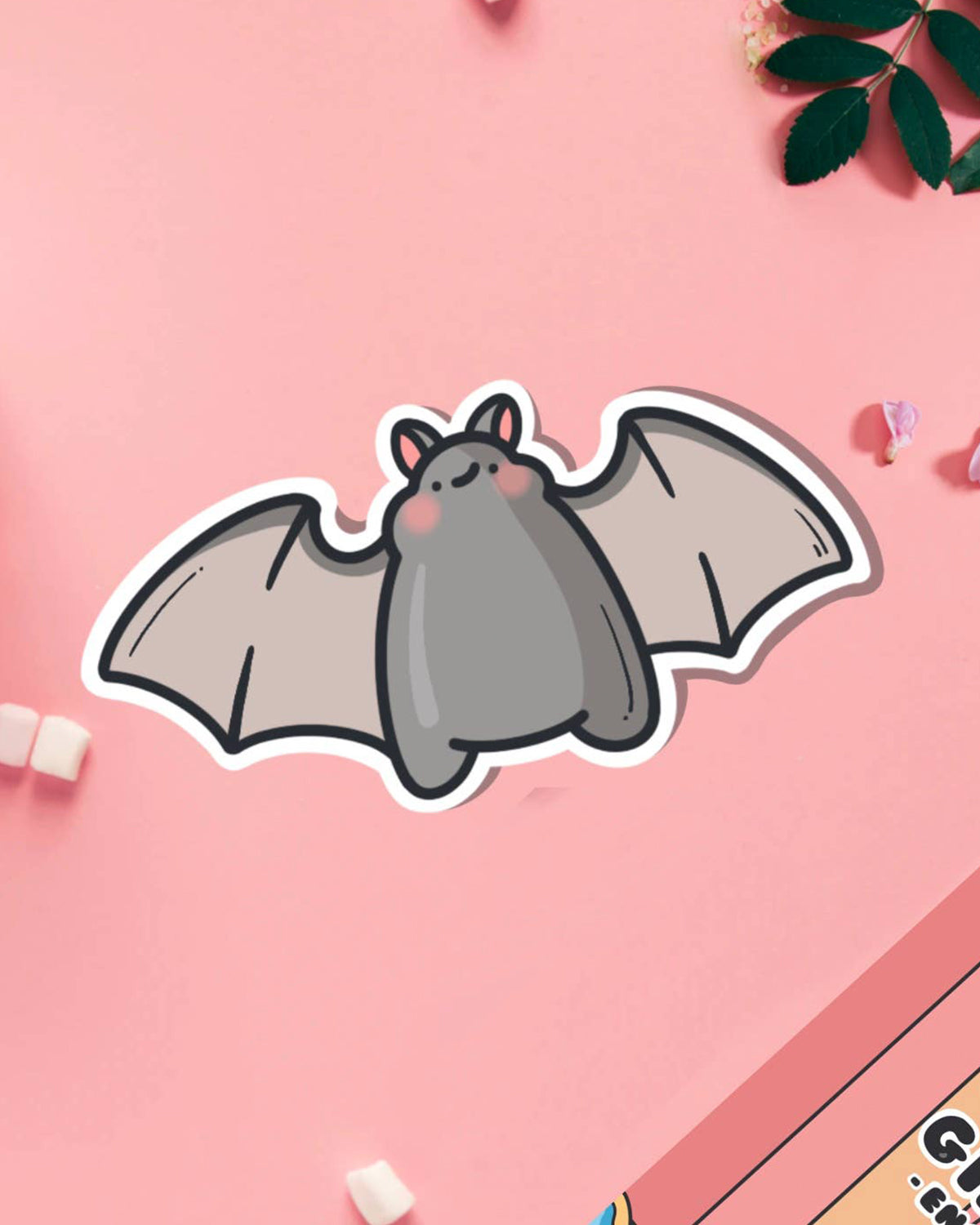 A cute vinyl sticker of a small grey cartoon bat with pink cheeks and a sweet expression, wings outstretched. Shown on a pink background with marshmallows and flowers. A waterproof, glossy sticker perfect for laptops, notebooks, or water bottles.
