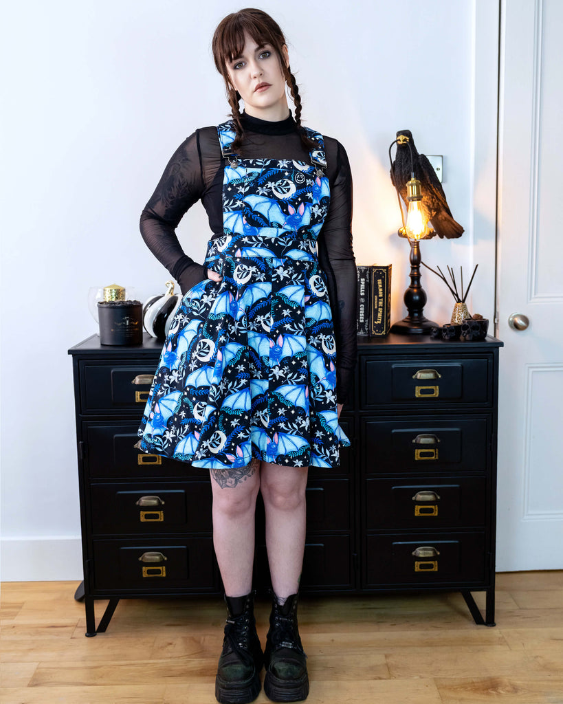 Alice is stood in front of a black dresser with creepy books and a crow lamp she is wearing a night flight flared pinafore and looks like Wednesday Addams. She has a black mesh top underneath Alice is looking into the camera with a straight face, She has her hand in the pocket.