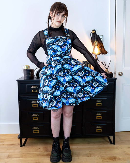 Alice is stood in front of a black dresser with creepy books and a crow lamp she is wearing a night flight flared pinafore and looks like Wednesday Addams. She has a black mesh top underneath Alice is looking into the camera with a straight face. She is holding one side of the pinafore flared out. 