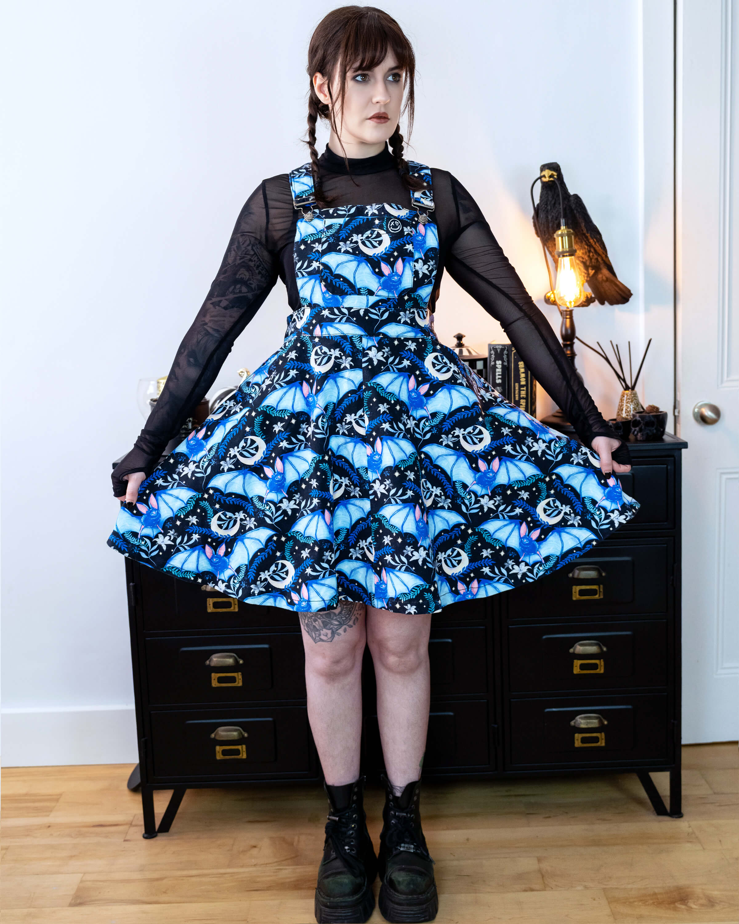 Alice is stood in front of a black dresser with creepy books and a crow lamp she is wearing a night flight flared pinafore and looks like Wednesday Addams. She has a black mesh top underneath Alice is looking off to the side and of the camera with a straight face and is holding out the skirt. 