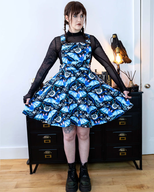 Alice is stood in front of a black dresser with creepy books and a crow lamp she is wearing a night flight flared pinafore and looks like Wednesday Addams. She has a black mesh top underneath Alice is looking off to the side and of the camera with a straight face and is holding out the skirt. 