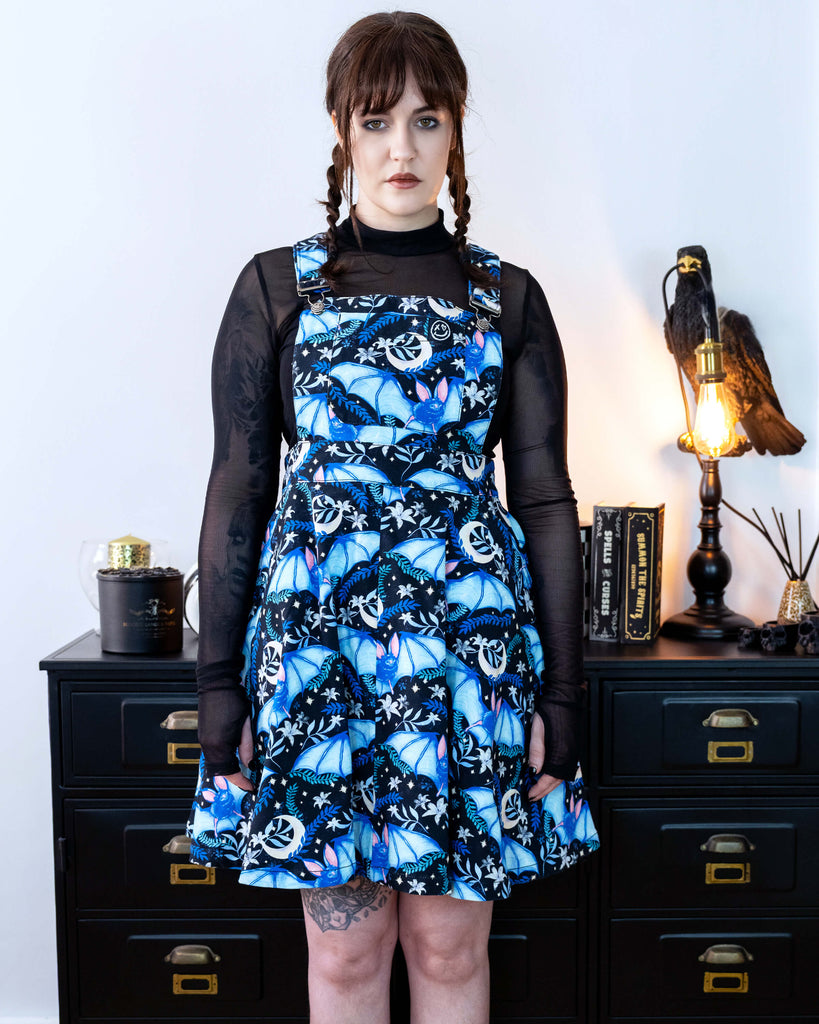 Alice is stood in front of a black dresser with creepy books and a crow lamp she is wearing a night flight flared pinafore and looks like Wednesday Addams. She has a black mesh top underneath Alice is looking into the camera with a straight face.