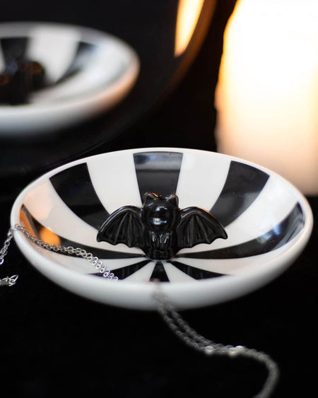 The striped bat trinket dish displayed on a black surface with a silver chain inside, illuminated by soft candlelight in the background.