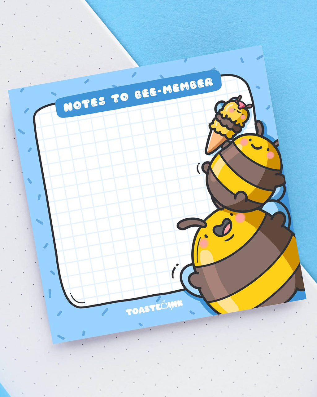 Square sticky note pad with a blue border and grid paper, featuring two chubby yellow and brown cartoon bumble bees stacking on top of each other to reach an ice cream. The top reads “Notes to Bee-member” in bold white text. Cute and practical stationery for daily reminders.