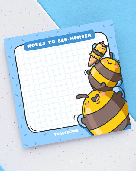Square sticky note pad with a blue border and grid paper, featuring two chubby yellow and brown cartoon bumble bees stacking on top of each other to reach an ice cream. The top reads “Notes to Bee-member” in bold white text. Cute and practical stationery for daily reminders.