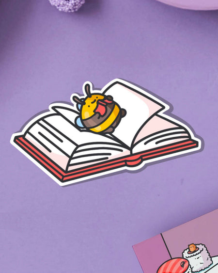 A cute vinyl sticker of a yellow and brown bumblebee lying on its back on top of an open book, tongue sticking out playfully. Displayed on a purple background with pink and blue elements. Glossy waterproof finish, ideal for book lovers and readers.