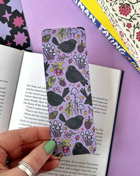  A hand with green nail polish holding a nature-themed bookmark featuring blackbirds, mushrooms, berries, and autumn leaves on a soft lavender background. An open book and colourful covers are in the background.
