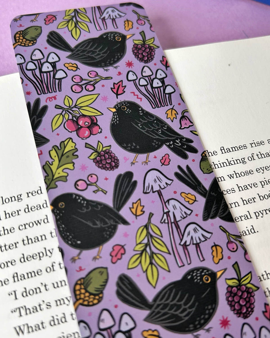 A detailed view of the blackbird design with clusters of berries, mushrooms, and leaves on a lavender background. The bookmark rests on an open book.