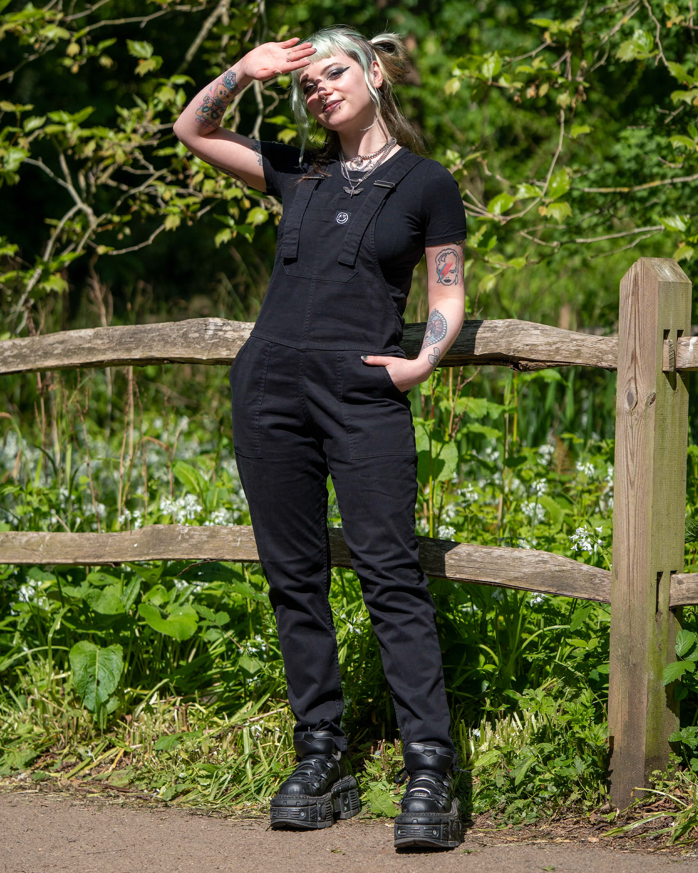 Model wearing Rebel Romance Jet Black Dungarees standing by a wooden fence, saluting toward the sun.