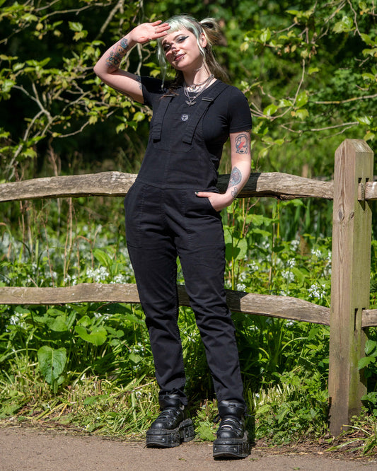 Model wearing Rebel Romance Jet Black Dungarees standing by a wooden fence, saluting toward the sun.