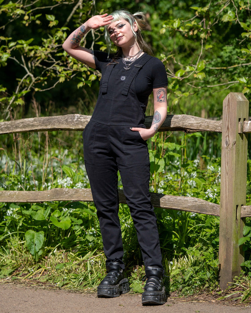 Model wearing Rebel Romance Jet Black Dungarees standing by a wooden fence, saluting toward the sun.