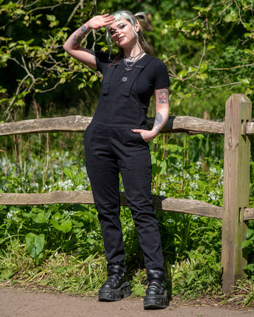 Model wearing Rebel Romance Jet Black Dungarees standing by a wooden fence, saluting toward the sun.
