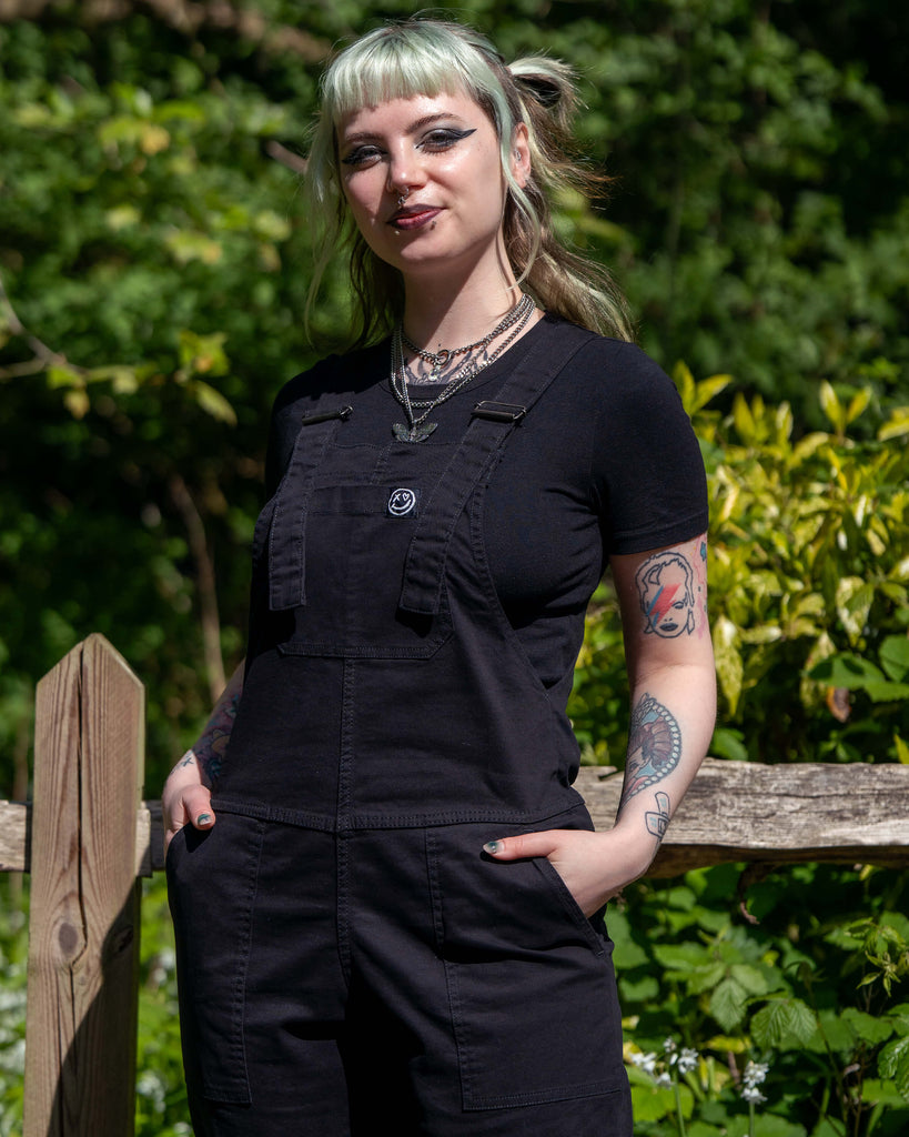 Portrait shot of model smiling in Rebel Romance Jet Black Dungarees paired with a black t-shirt.