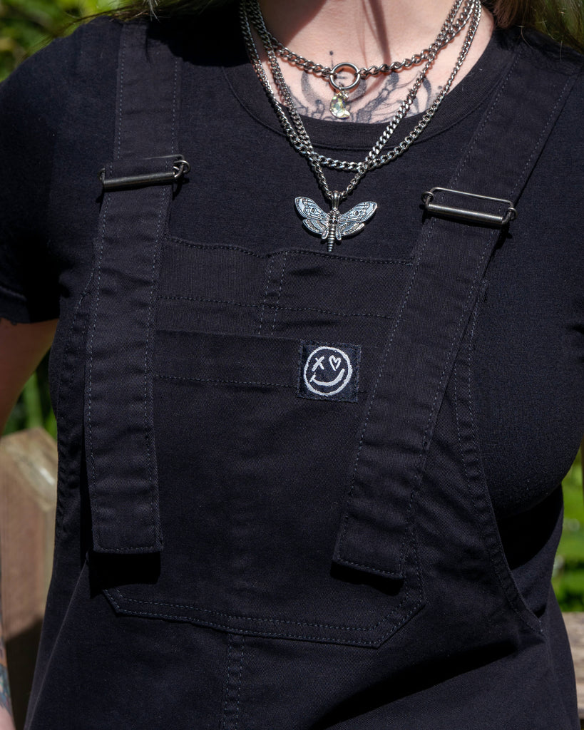 Close-up of the front bib and strap detail of Rebel Romance Jet Black Dungarees with smiley logo patch.