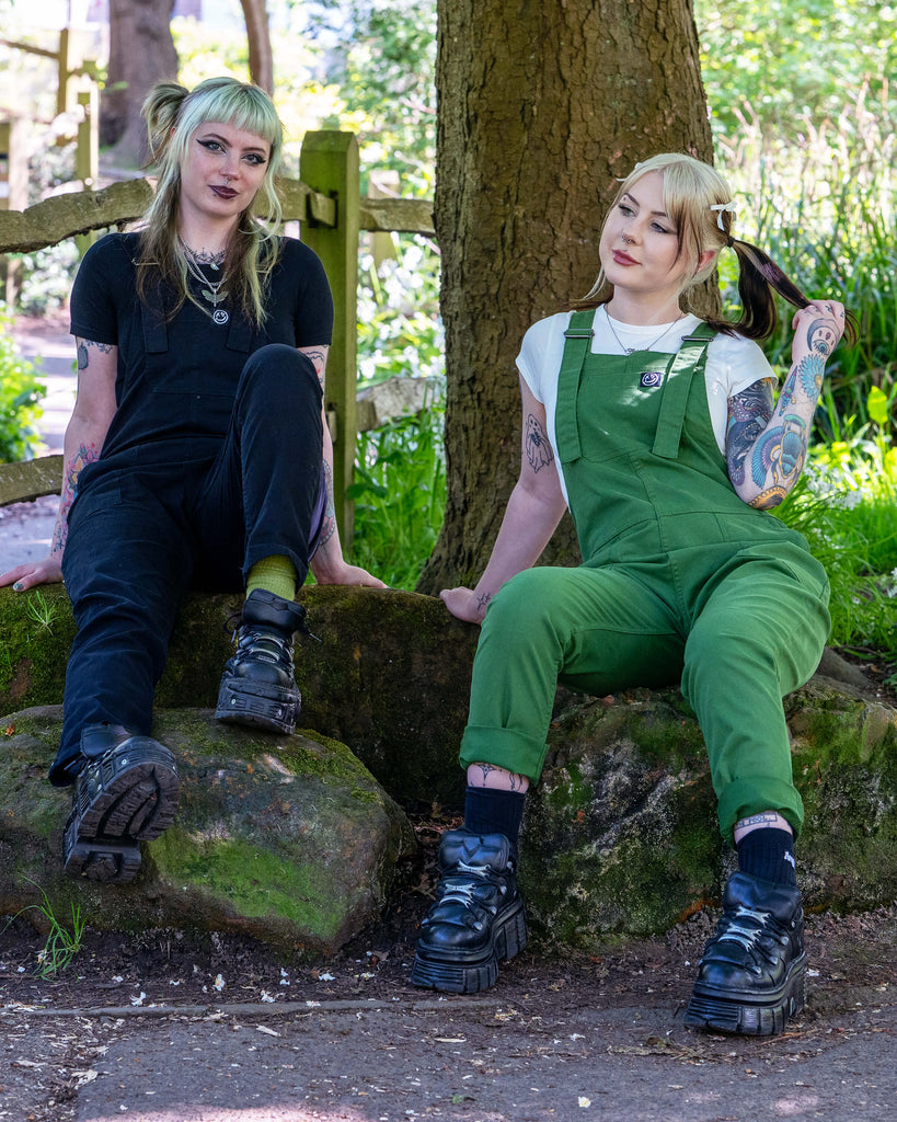 Two models sitting outdoors on mossy rocks, one in black dungarees and one in green, smiling and relaxed.