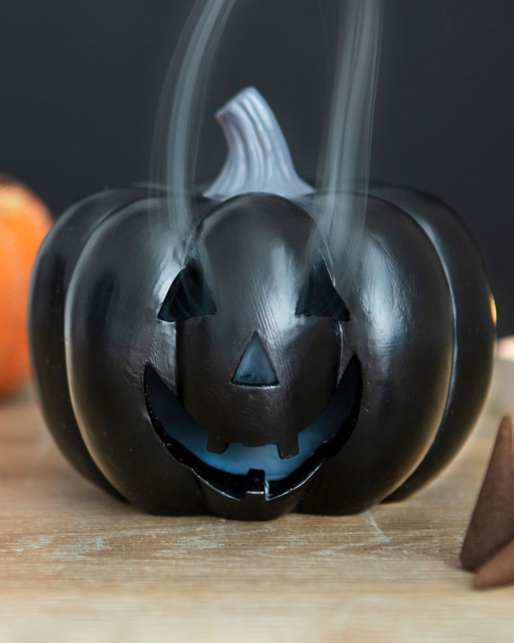 Close-up of the black pumpkin incense holder in use, with smoke curling from its carved features, placed on a wooden surface with a blurred background.