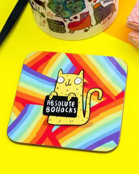 Colourful square coaster with a yellow cartoon cat holding a sign that reads "Absolute Bollocks" on a vibrant rainbow-striped background. Designed by Katie Abey.