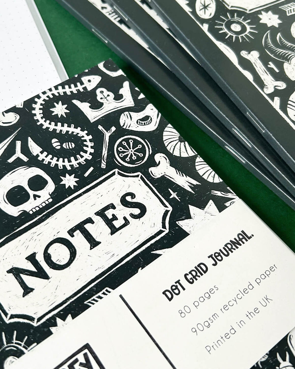 Close-up photo of a black and white dot grid journal with a hand-illustrated cover featuring skulls, bones, stars, and crowns, inspired by archaeology, folklore, and witchy aesthetics. The notebook has a white band with the words “Notes” and details of the journal printed on it.