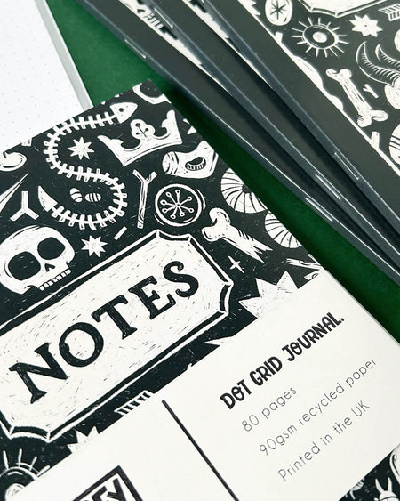 Close-up photo of a black and white dot grid journal with a hand-illustrated cover featuring skulls, bones, stars, and crowns, inspired by archaeology, folklore, and witchy aesthetics. The notebook has a white band with the words “Notes” and details of the journal printed on it.