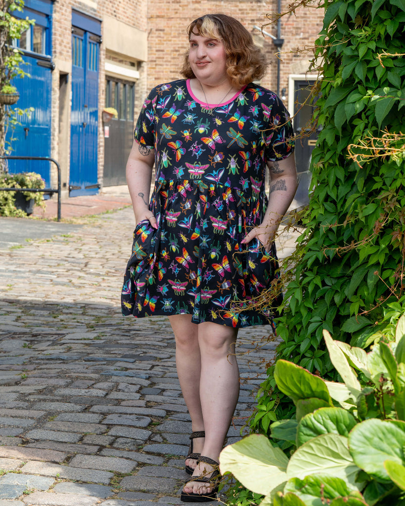 Maddie wears the Rebel Romance OUTLET PRIDE Bugs Stretch Smock Dress on a cobbled street. The black dress is covered in bright rainbow insect prints and has pockets, giving a bold alternative fashion vibe from this UK ethical brand.