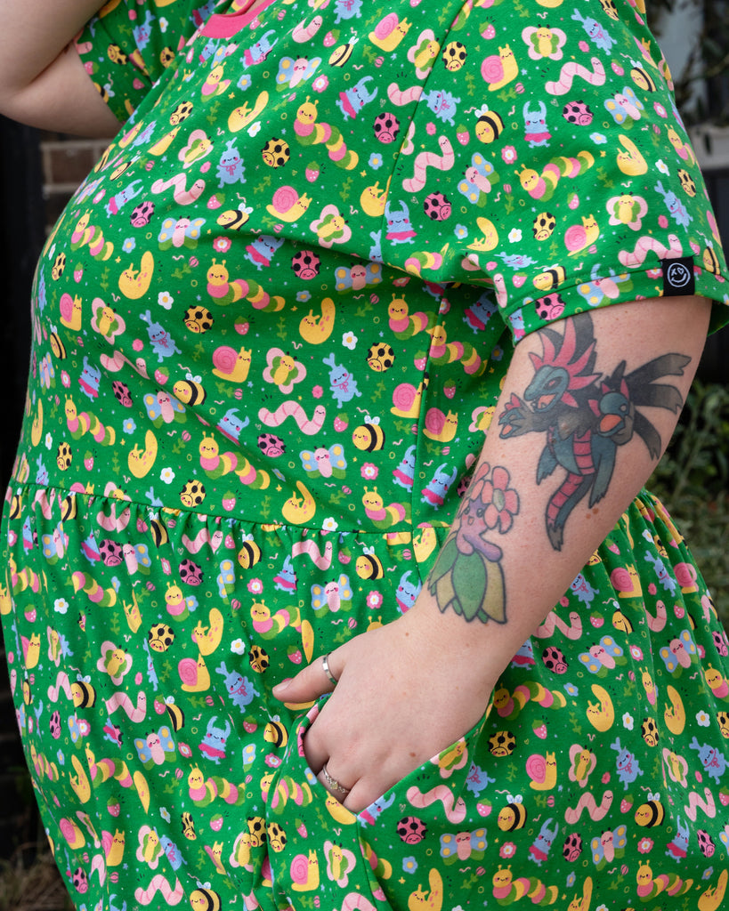 Close-up of a hand tucked into the pocket of a green dress decorated with a playful pastel bug print. The pattern includes smiling caterpillars, snails, butterflies, ladybirds, and flowers against a bright green background.